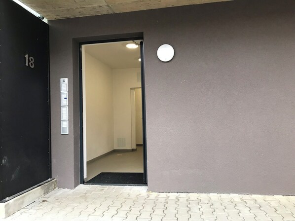 An entrance to a building with a black door marked with the number 18, a set of four control panels on the wall, and a circular light fixture above the door.