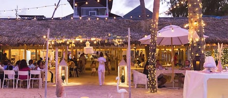 Guests can dine al fresco under a thatched roof, surrounded by palm trees and string lights.