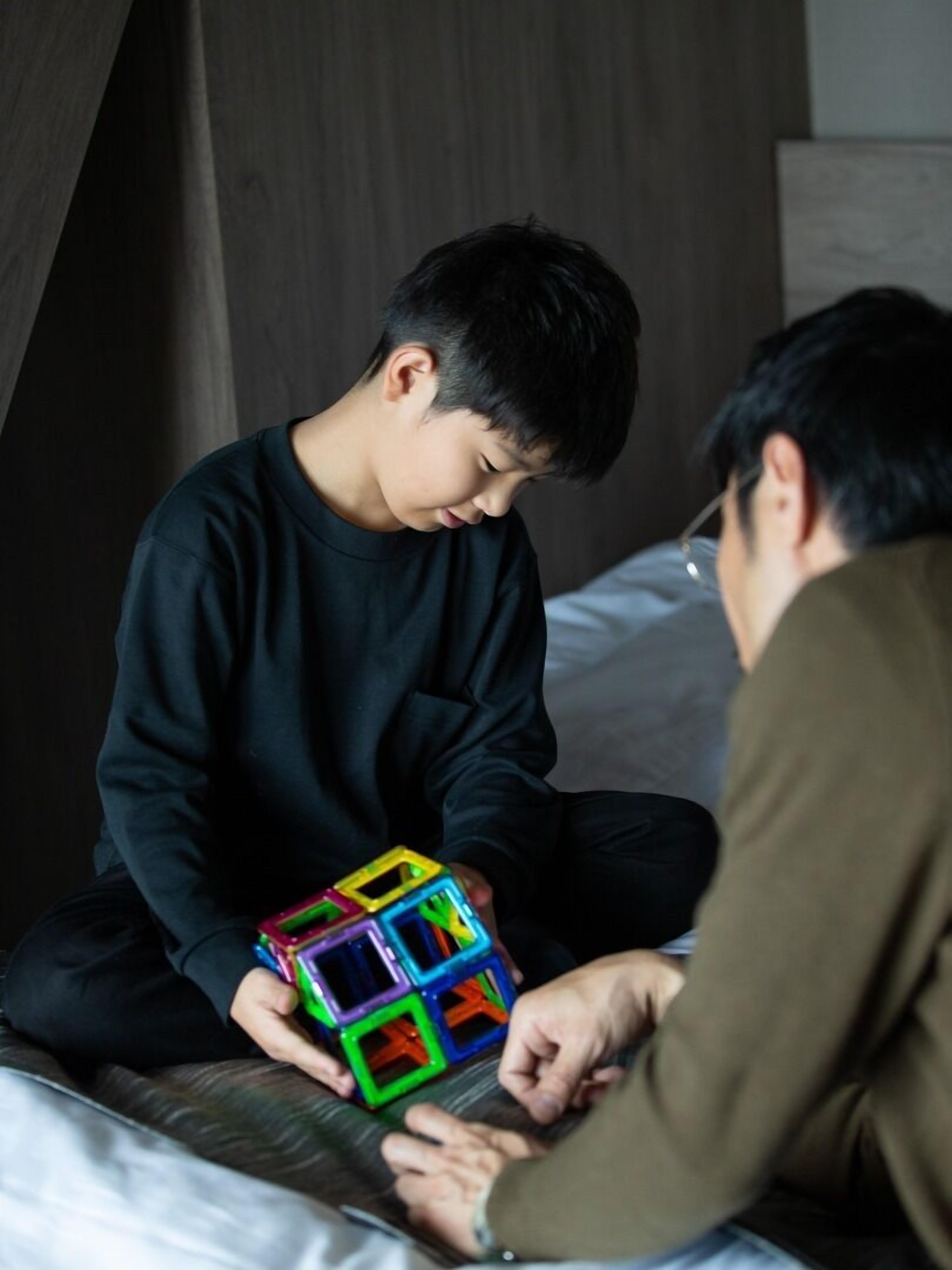 Dos personas sentadas en una cama con cabecera de madera, una sosteniendo un cubo de Rubik colorido.