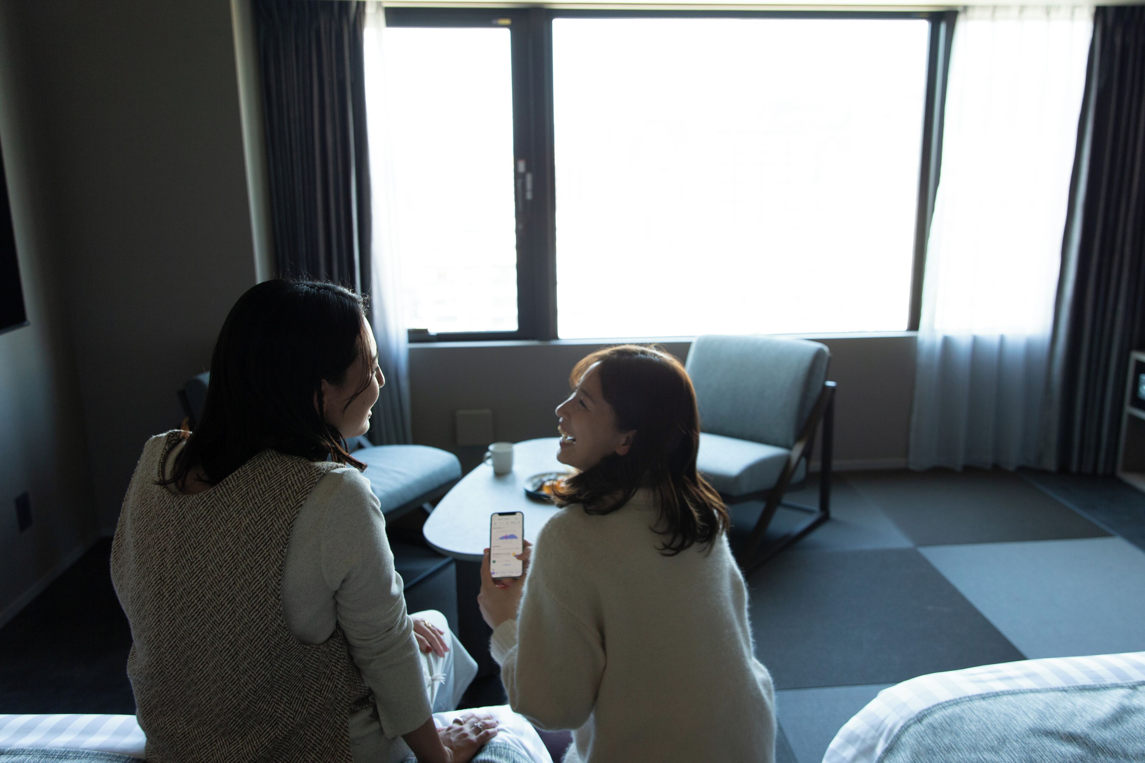 Two people sitting on a bed in a hotel room with a window, a small table, and a chair.