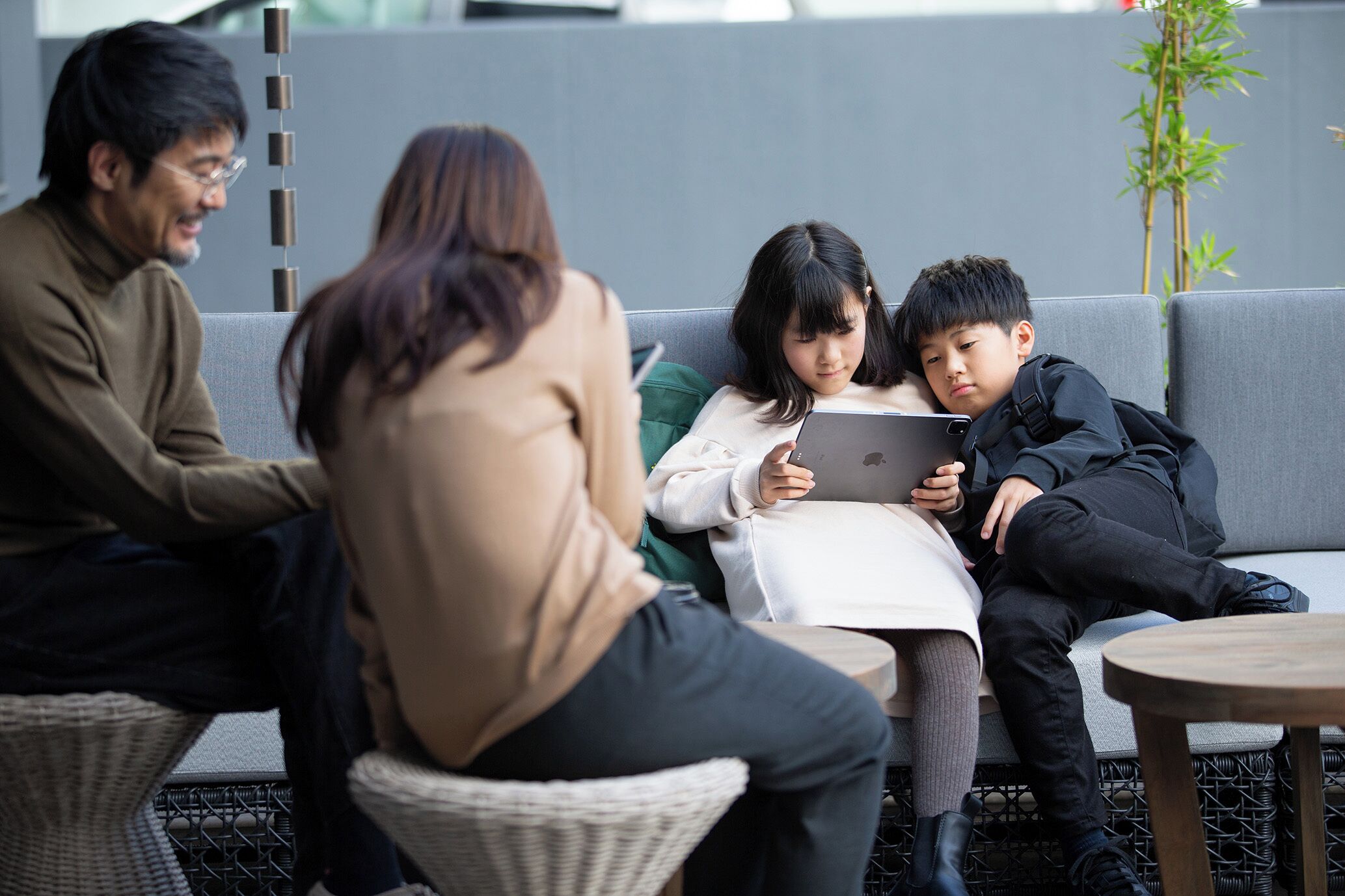 Eine Gruppe von Menschen sitzt auf einer grauen Couch und Sesseln in einem modernen Innenraum, mit einem Korbsessel und einem kleinen runden Tisch.