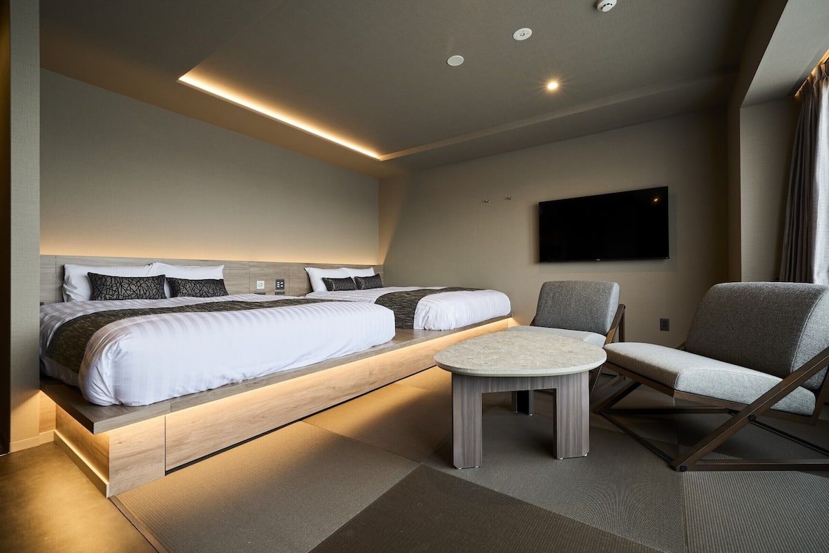 A modern hotel room with a large bed, a TV mounted on the wall, and a seating area with a table and chairs.