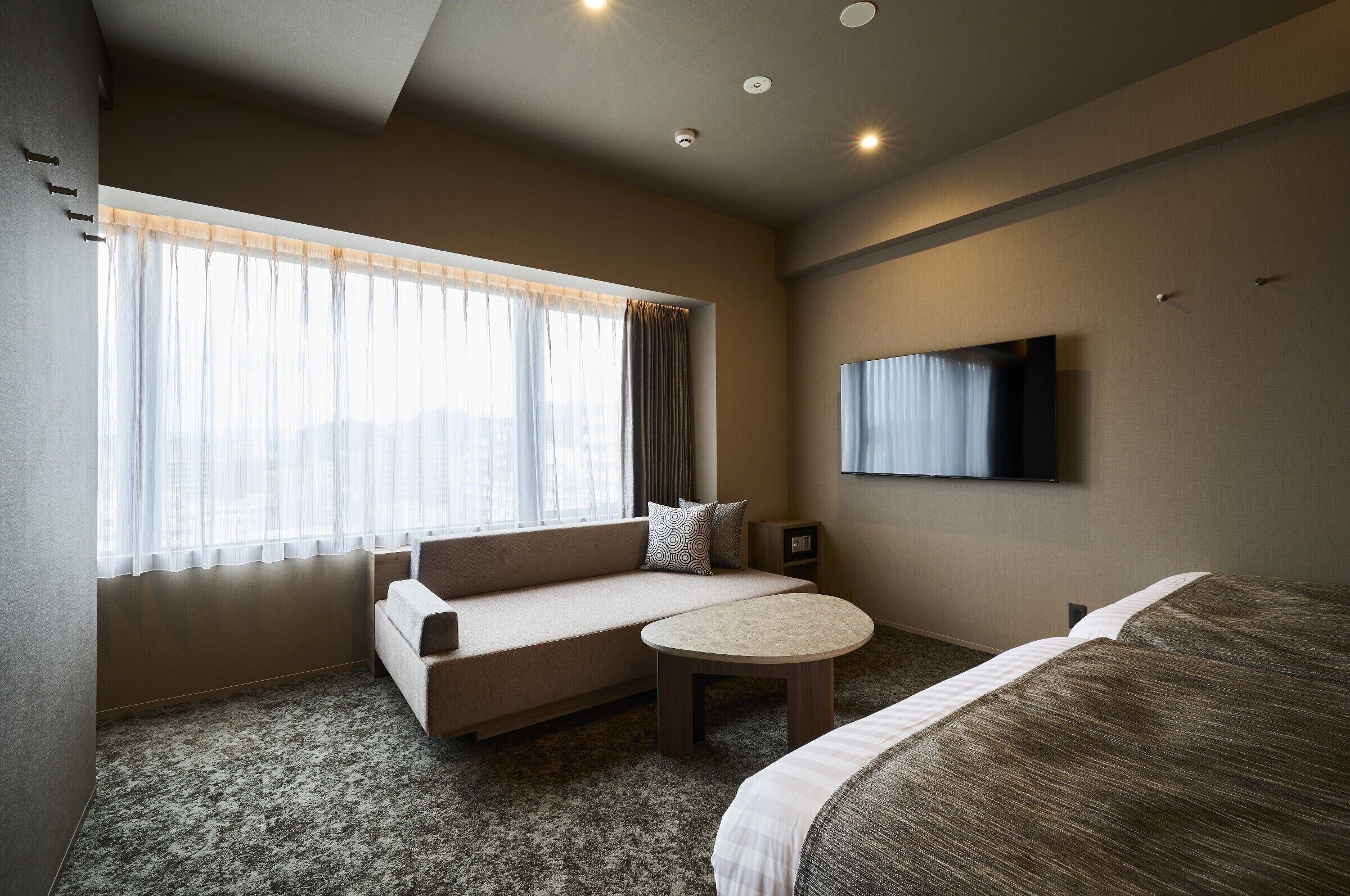 A hotel room with a sofa bed, a small round table, a TV mounted on the wall, and a large window with curtains.