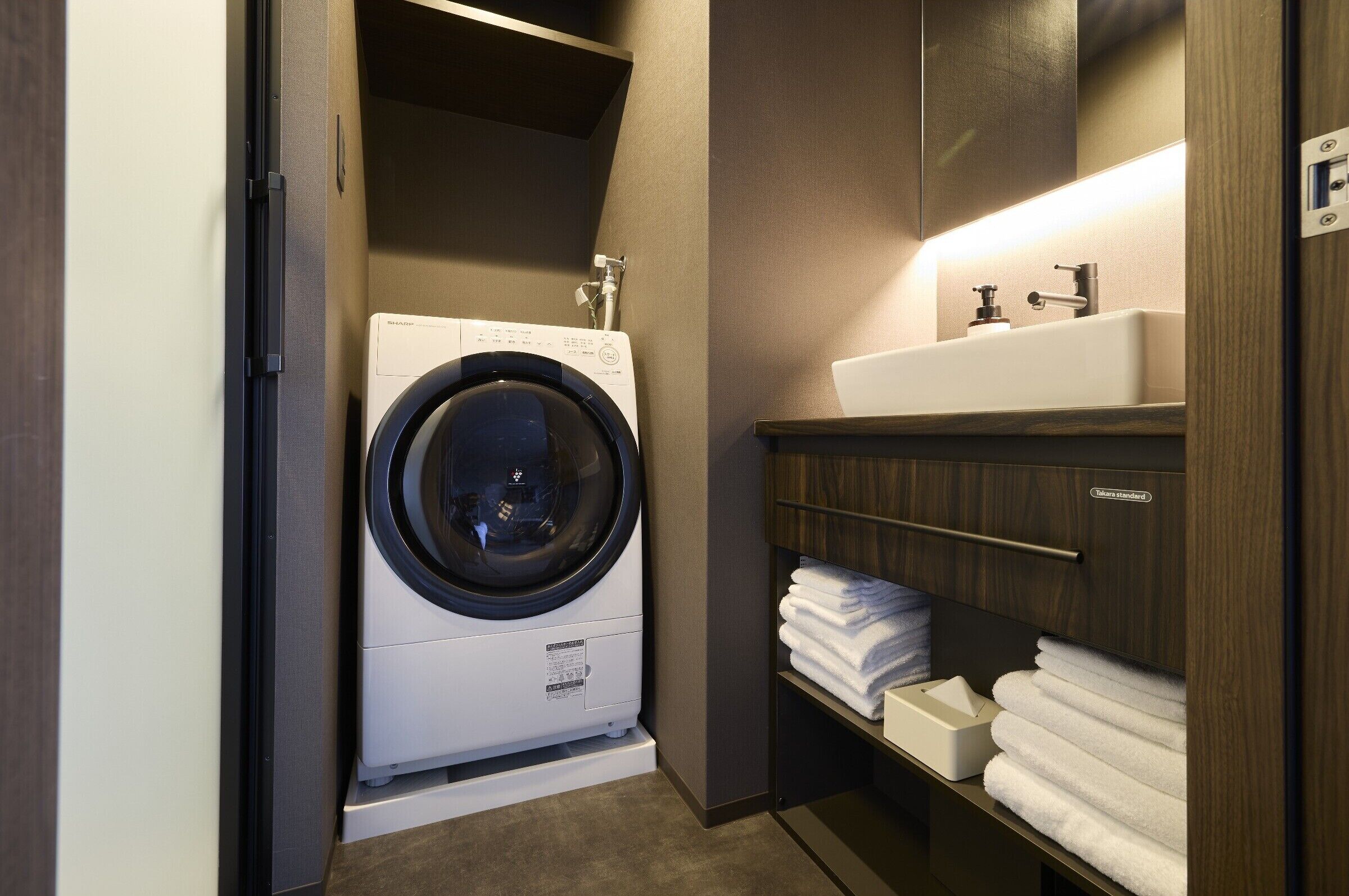 Een kleine hotelbadkamer met een voorlader wasmachine, een wastafel en een plank met keurig opgevouwen handdoeken.