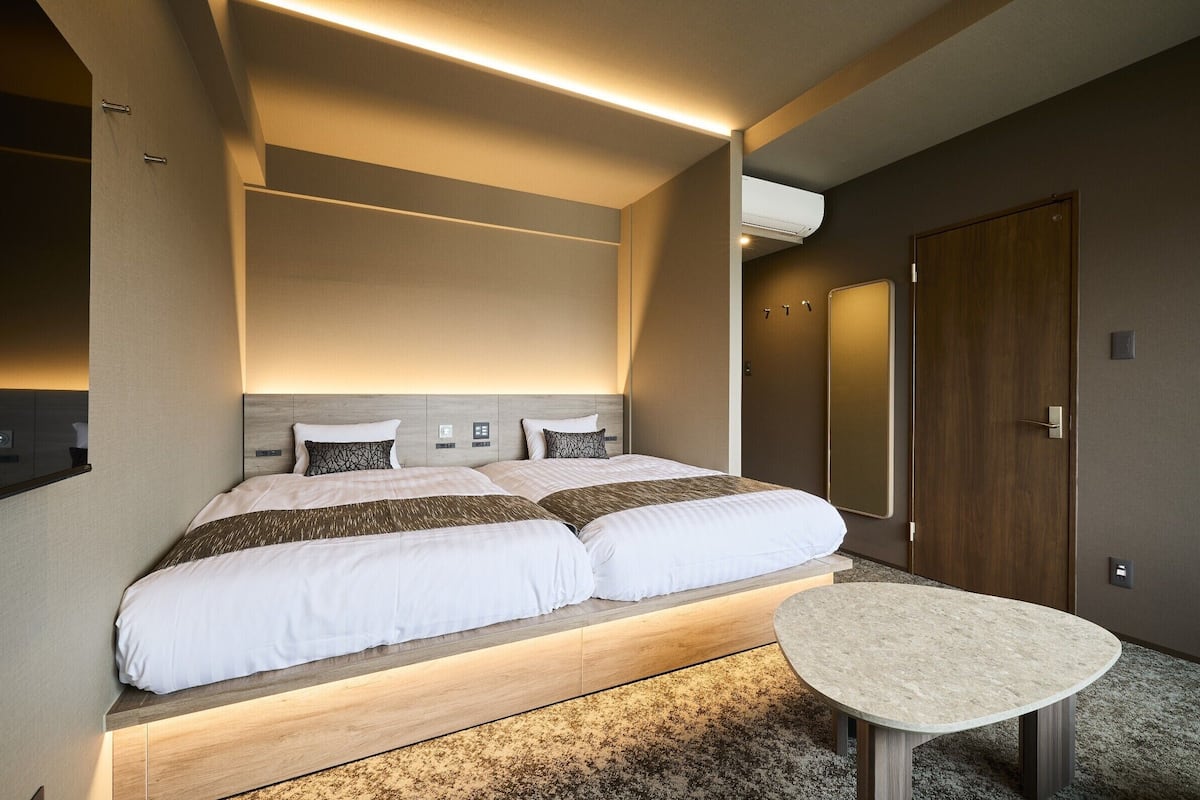 A modern hotel room with a large bed, bedside tables, a small round table, and a built-in headboard.