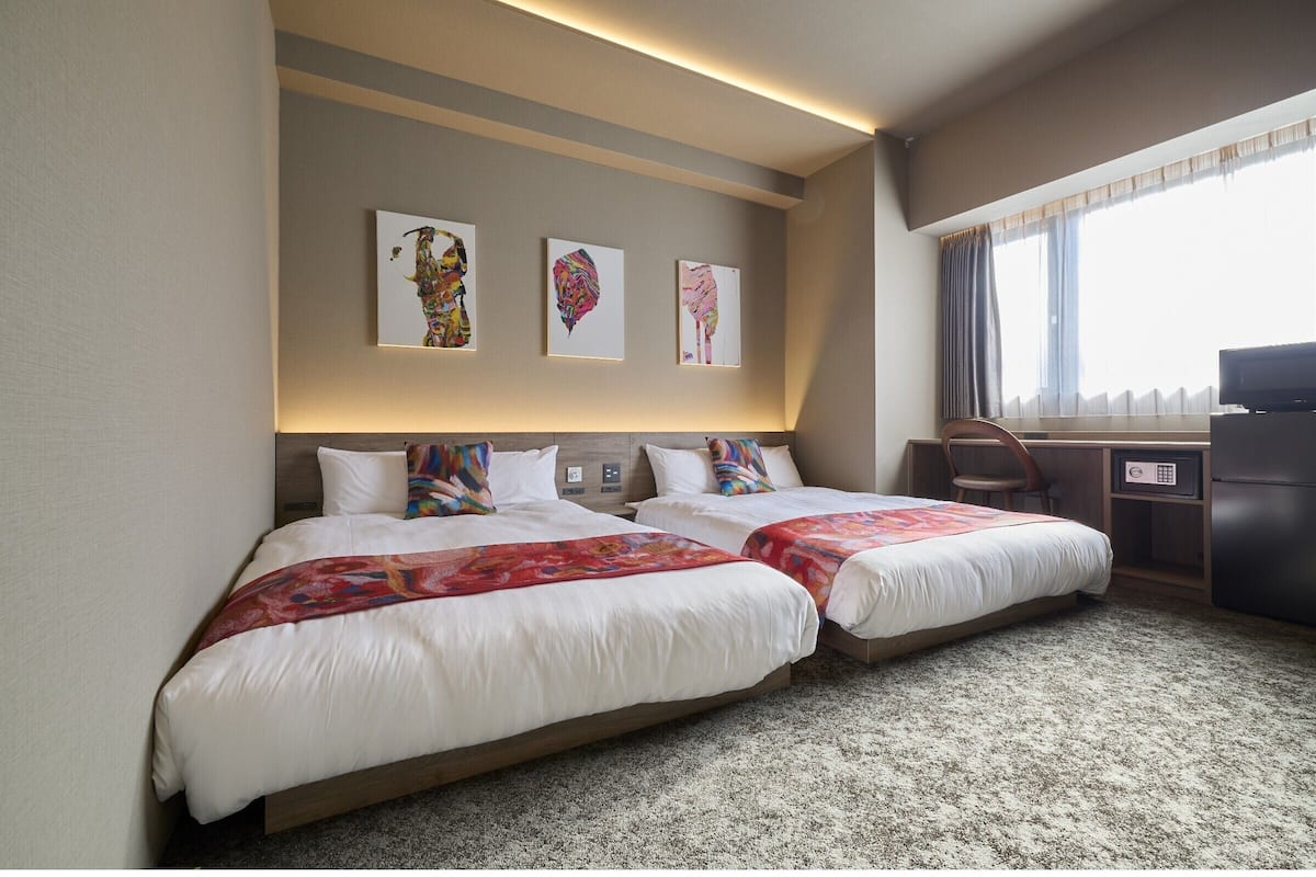 A hotel room with two beds, a desk, a chair, and artwork on the wall.