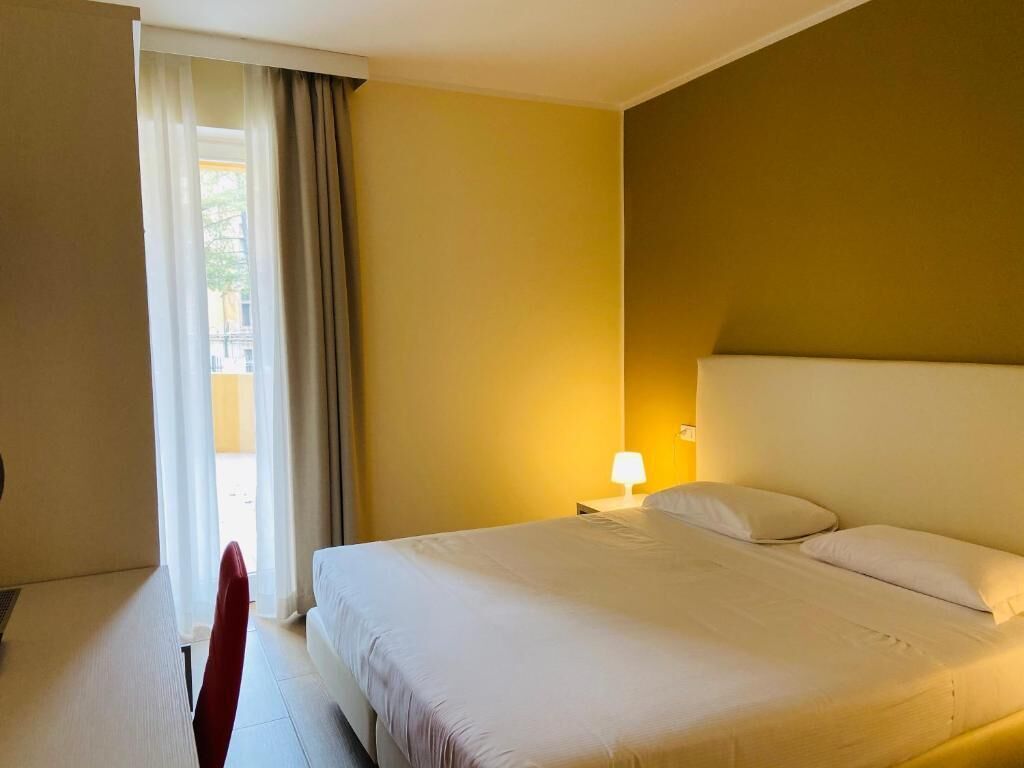 A hotel room with a bed, a nightstand, a lamp, and a window with curtains.