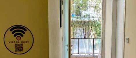 Un couloir avec un mur jaune, un symbole Wi-Fi et une porte donnant sur un balcon avec vue sur des arbres et un immeuble.