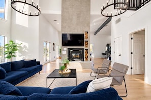 A spacious living area with a large sofa set, a coffee table, and a fireplace. There is a television mounted on the wall and a staircase leading to an upper level. - Modern Home - 1 Acre Lot w/ Pool, Patios, Jacuzzi (Denton)