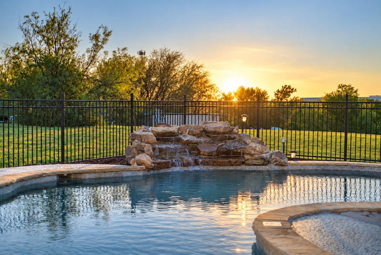 A swimming pool with a rock waterfall feature, surrounded by a black metal fence and a grassy area with trees.