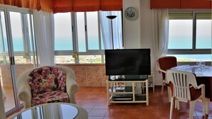 A living room with a TV, a floral armchair, a dining table with chairs, and a view of the sea through large windows. - Penthouse-apartment mit 140 qm - Direkter Merresblick (Monte Faro)
