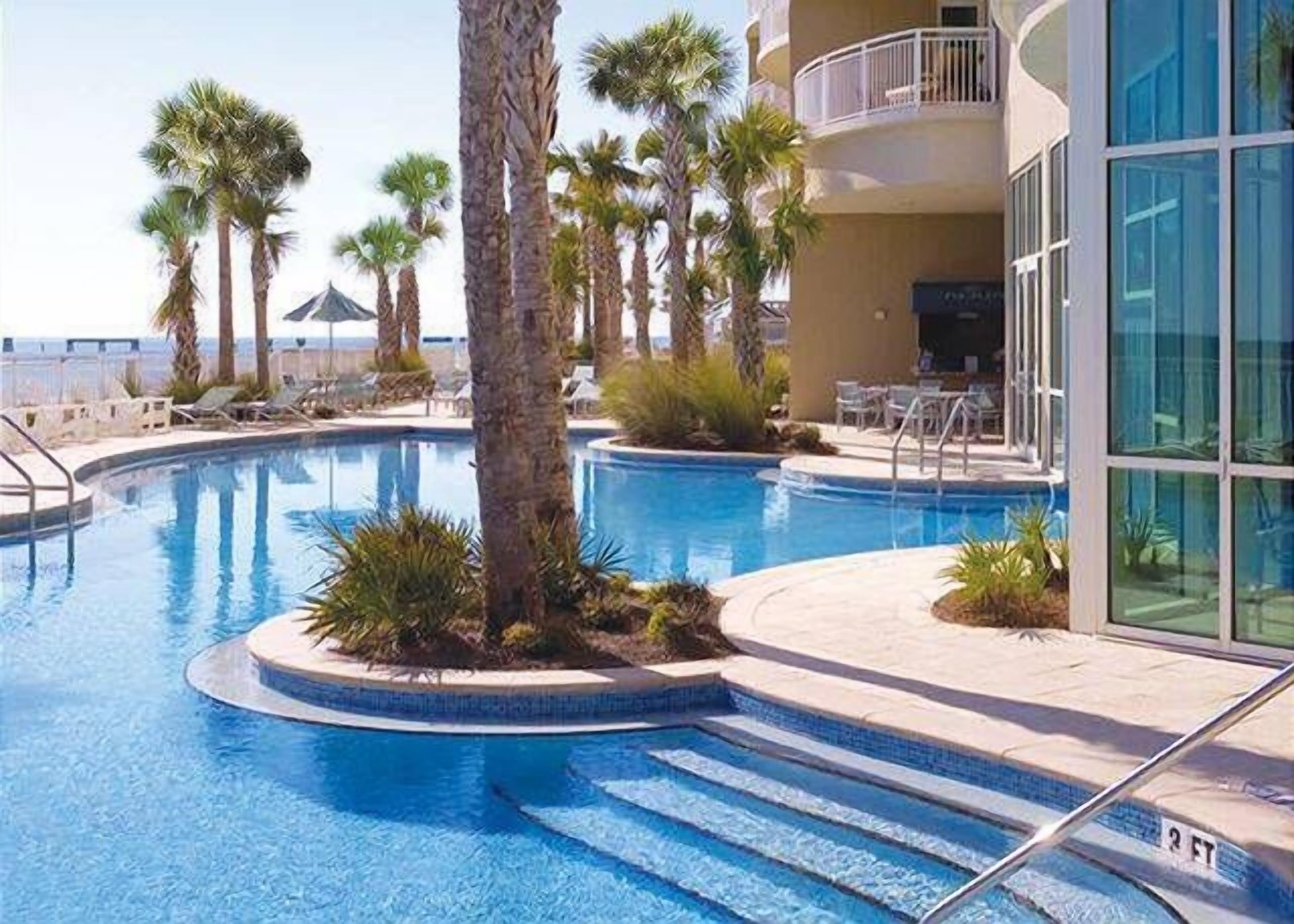 A swimming pool with palm trees, a sun terrace, and a building with a balcony.