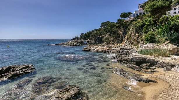 A coastal landscape with clear water, rocky shore, and a building on a cliff.