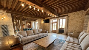 A spacious living room with a high ceiling and exposed wooden beams, a stone wall, and a large window. - La Bastide De Chassagne-montrachet (Chassagne-Montrachet)