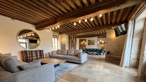 A spacious living room with a large sofa, a coffee table, and a stone fireplace. There is a stone wall and a wooden ceiling with exposed beams. - La Bastide De Chassagne-montrachet (Chassagne-Montrachet)