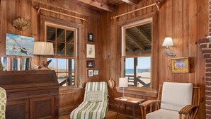 A wooden cabin interior with large windows, a piano, a striped armchair, and a wicker chair. - Fred Wood Cottage:Oceanfront, historic, Old Nags Head Style cottage, wide decks! (Nags Head)