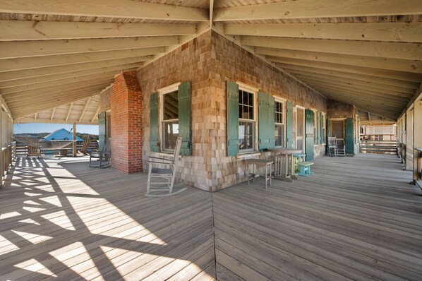 A covered wooden deck with a brick wall and green shutters, featuring outdoor seating and a view of the landscape.