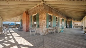 A covered wooden deck with a brick wall and green shutters, featuring outdoor seating and a view of the landscape. - Fred Wood Cottage:Oceanfront, historic, Old Nags Head Style cottage, wide decks! (Nags Head)