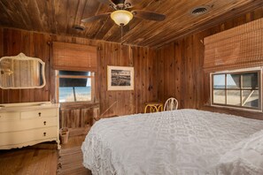 A bedroom with wooden walls, a ceiling fan, a bed with a white bedspread, a dresser, a chair, and a window with a view of the beach.