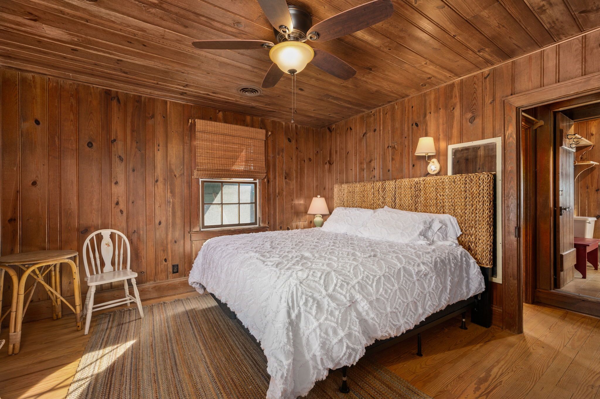 Un dormitorio con paneles de madera, una cama, una silla, una mesa y un ventilador de techo.