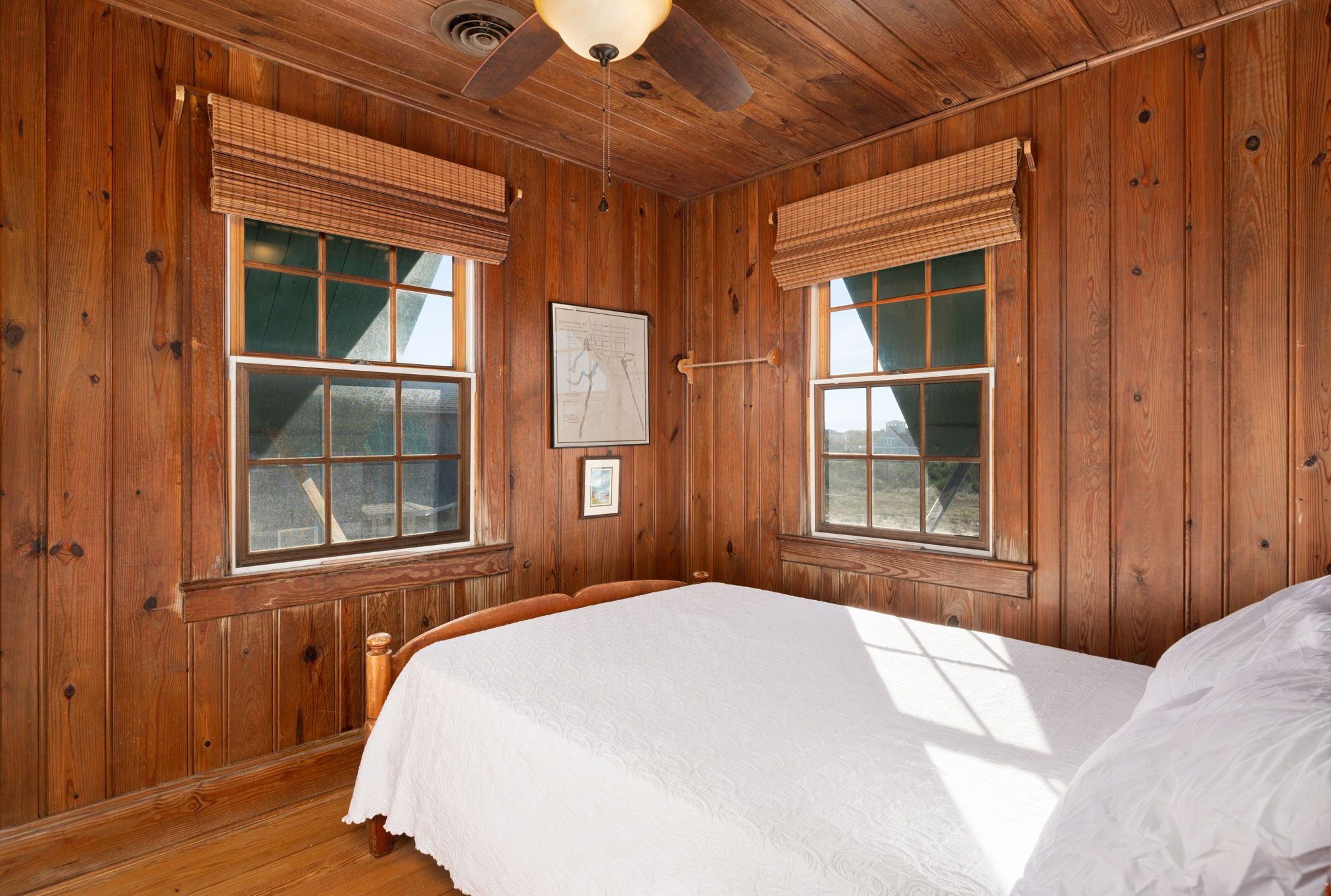 A wooden-paneled room with a bed, two windows with blinds, a ceiling fan, and a framed picture on the wall.