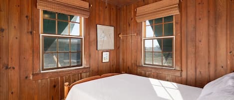 A wooden-paneled room with a bed, two windows with blinds, a ceiling fan, and a framed picture on the wall.