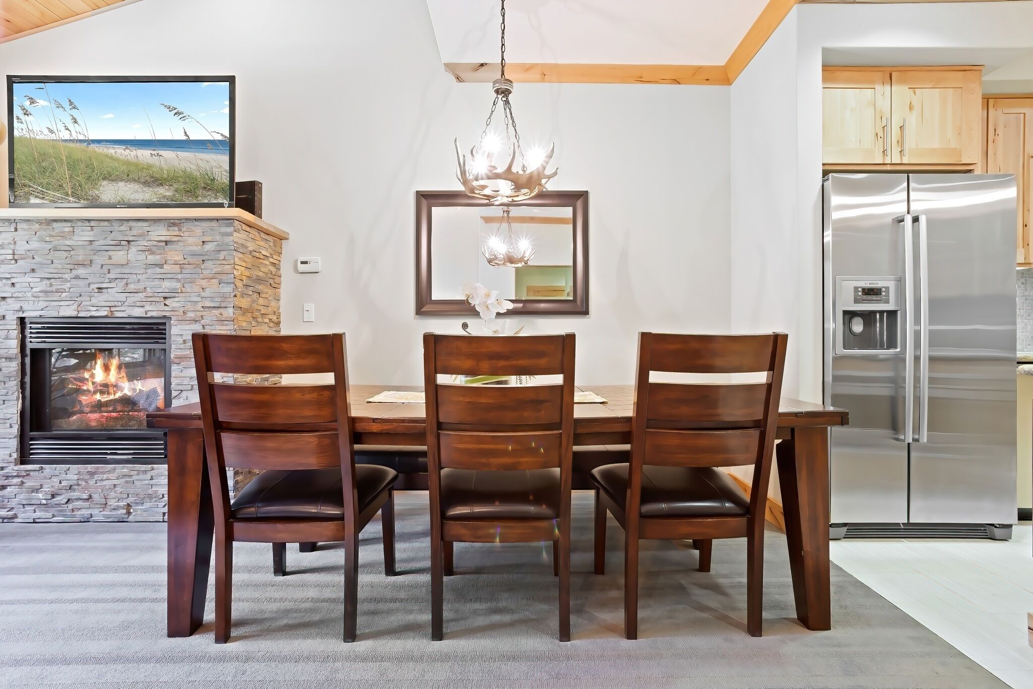 A dining area with a stone fireplace, a chandelier, and a modern kitchen with a stainless steel refrigerator.