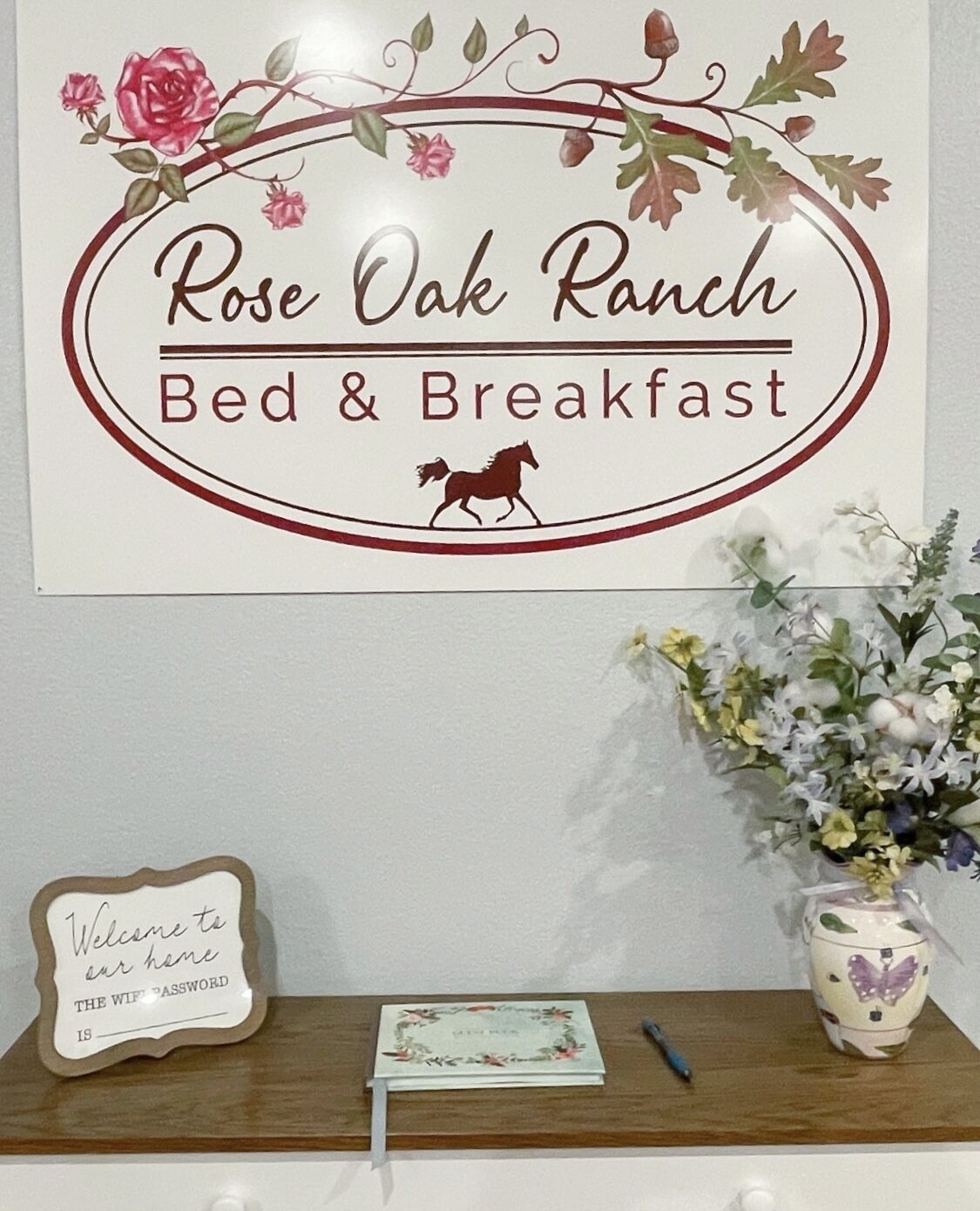 Schild für Rose Oak Ranch Bed & Breakfast mit einer Pferde-Silhouette, einem Willkommensschild, einem Notizblock und einer Vase mit Blumen.