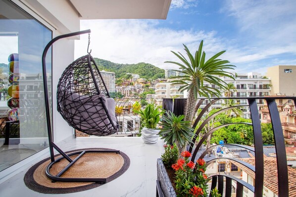 A balcony with a hanging chair, potted plants, and a view of a hillside town.