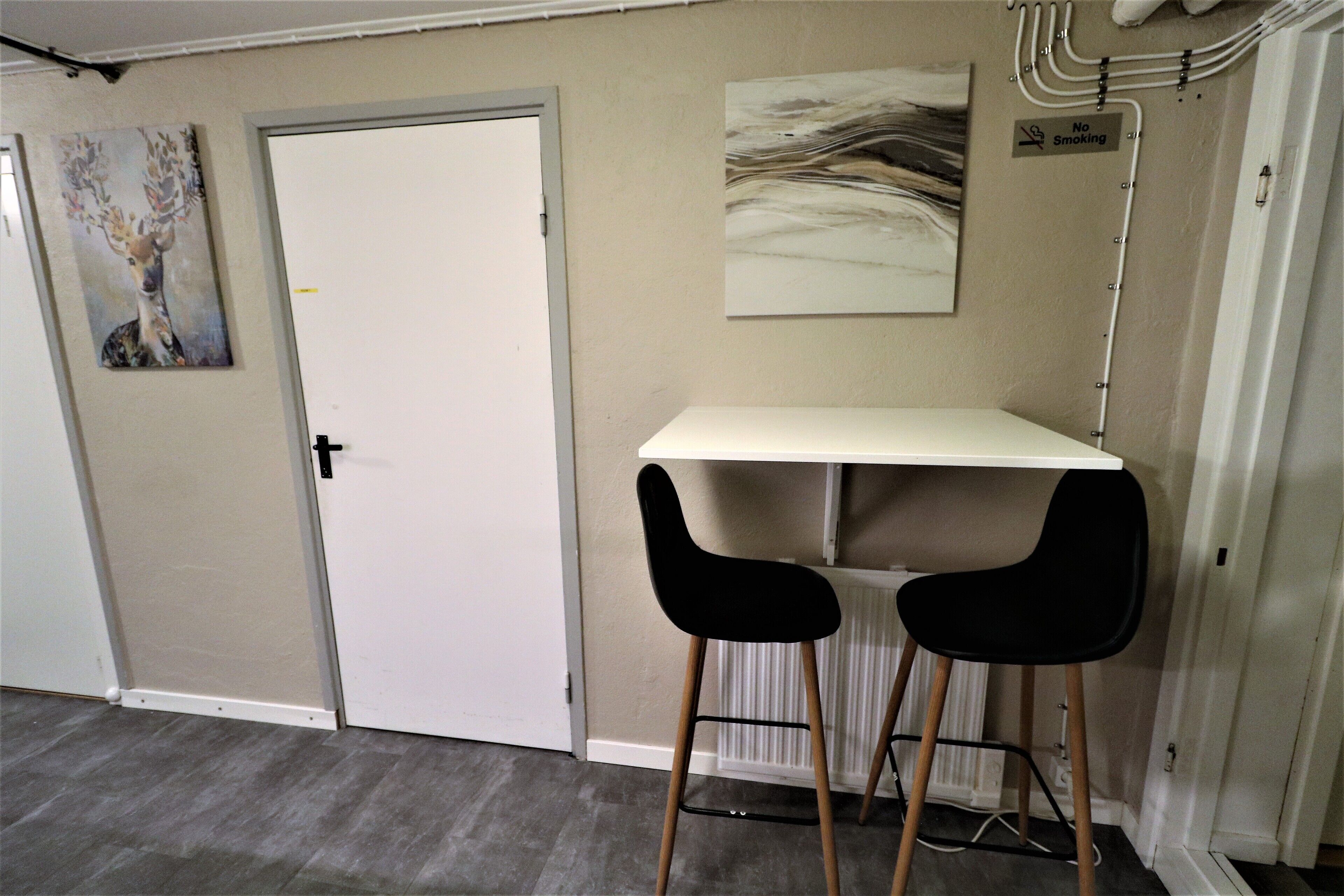 A room with a white door, a white desk, two black chairs, and two wall-mounted artworks.