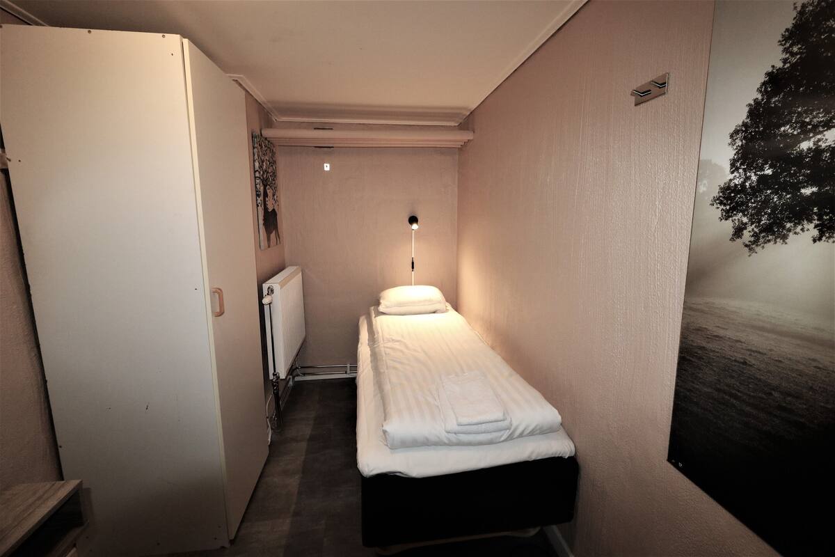 A narrow hallway leading to a single bed with a headboard, a radiator, and a wall-mounted light fixture.