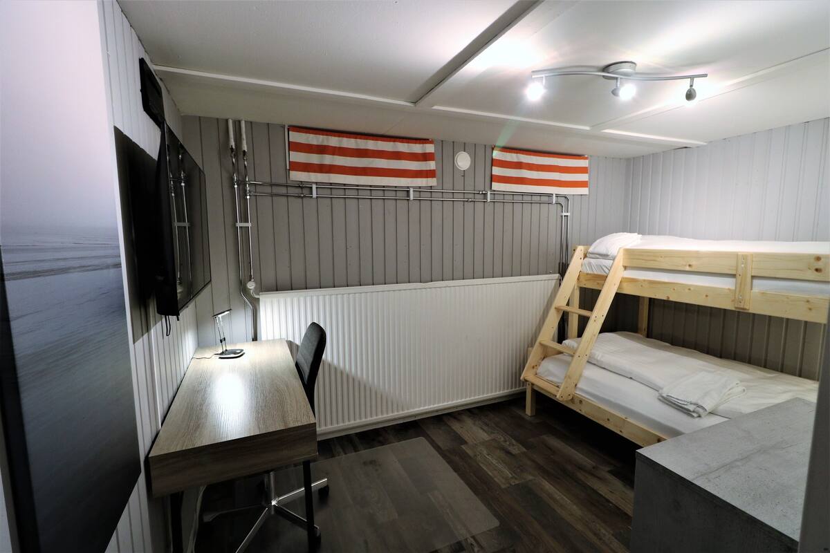 A compact room with a bunk bed, a desk with a chair, a television mounted on the wall, and a radiator.