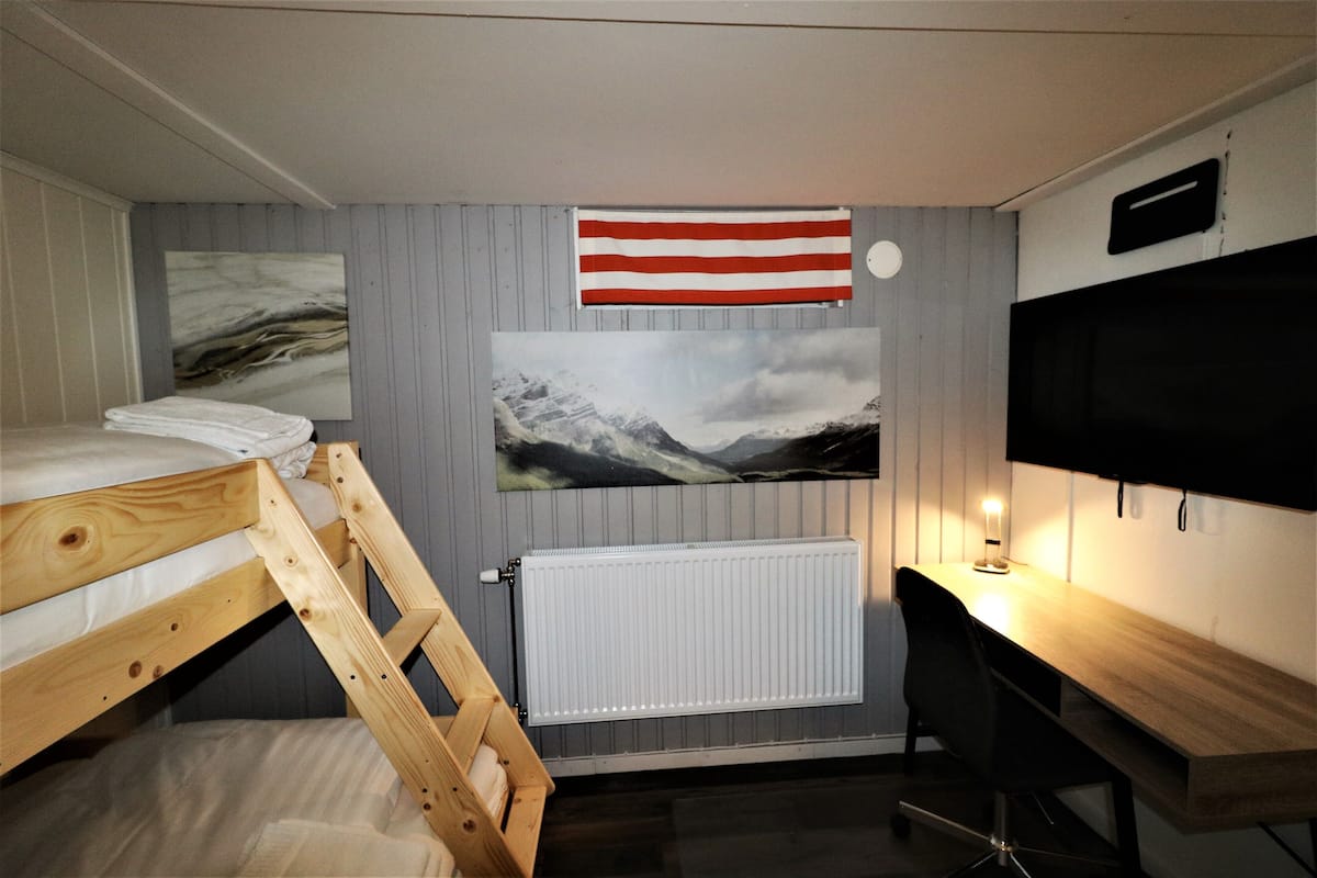 A bedroom with a bunk bed, a desk with a chair, a mounted TV, and a painting of a mountain landscape.