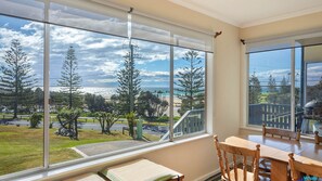 Enjoy expansive views of the coastal landscape from the large window, with a dining area featuring a table and chairs.