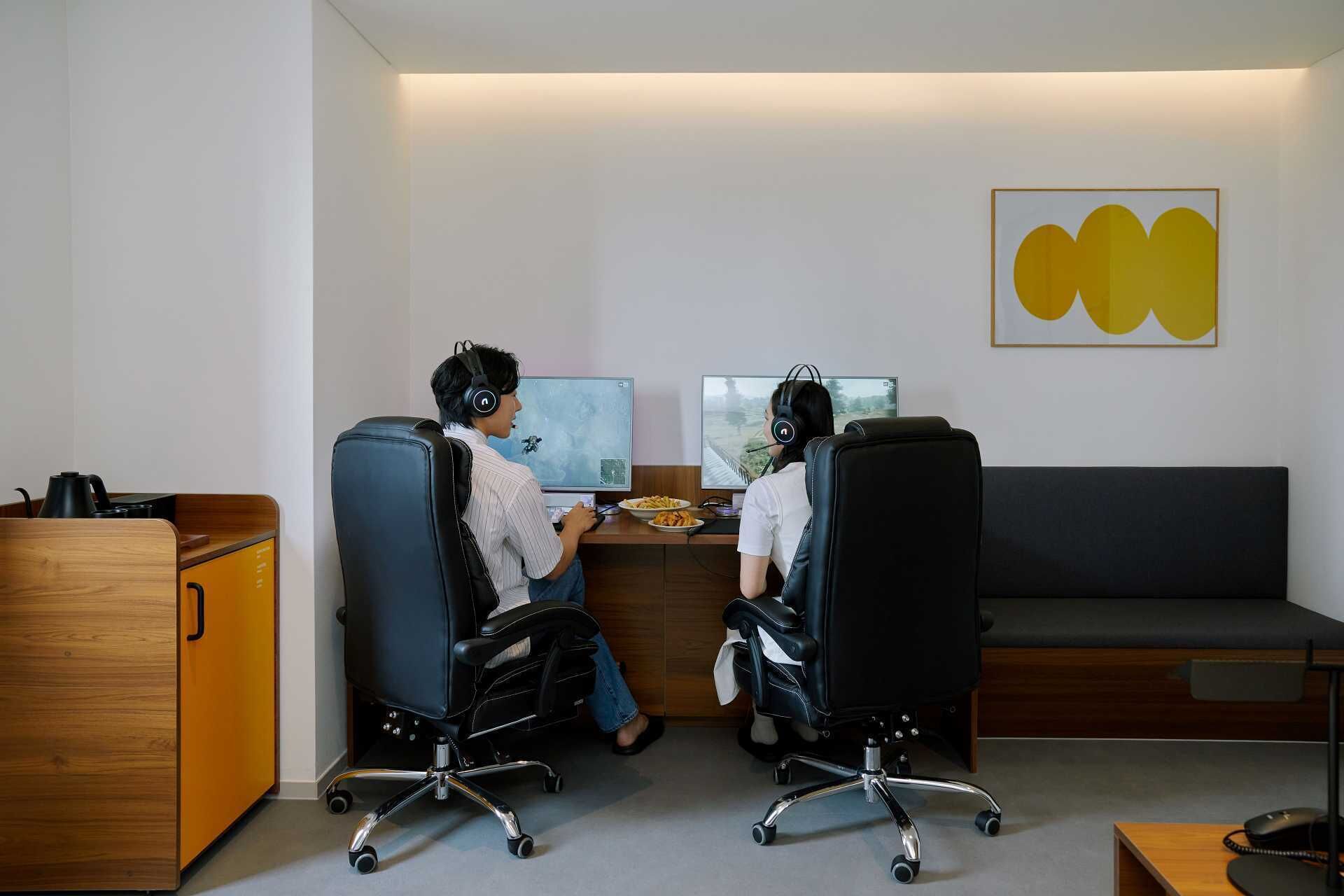 Two individuals seated at desks with computers, in a room with a wooden cabinet and a gray couch.