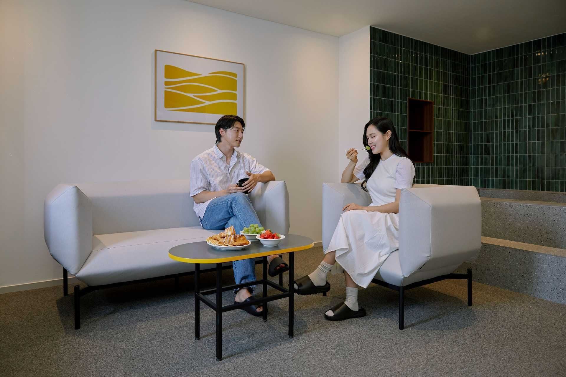 Dos personas sentadas en sofás modernos en un vestíbulo, con una pequeña mesa entre ellas.