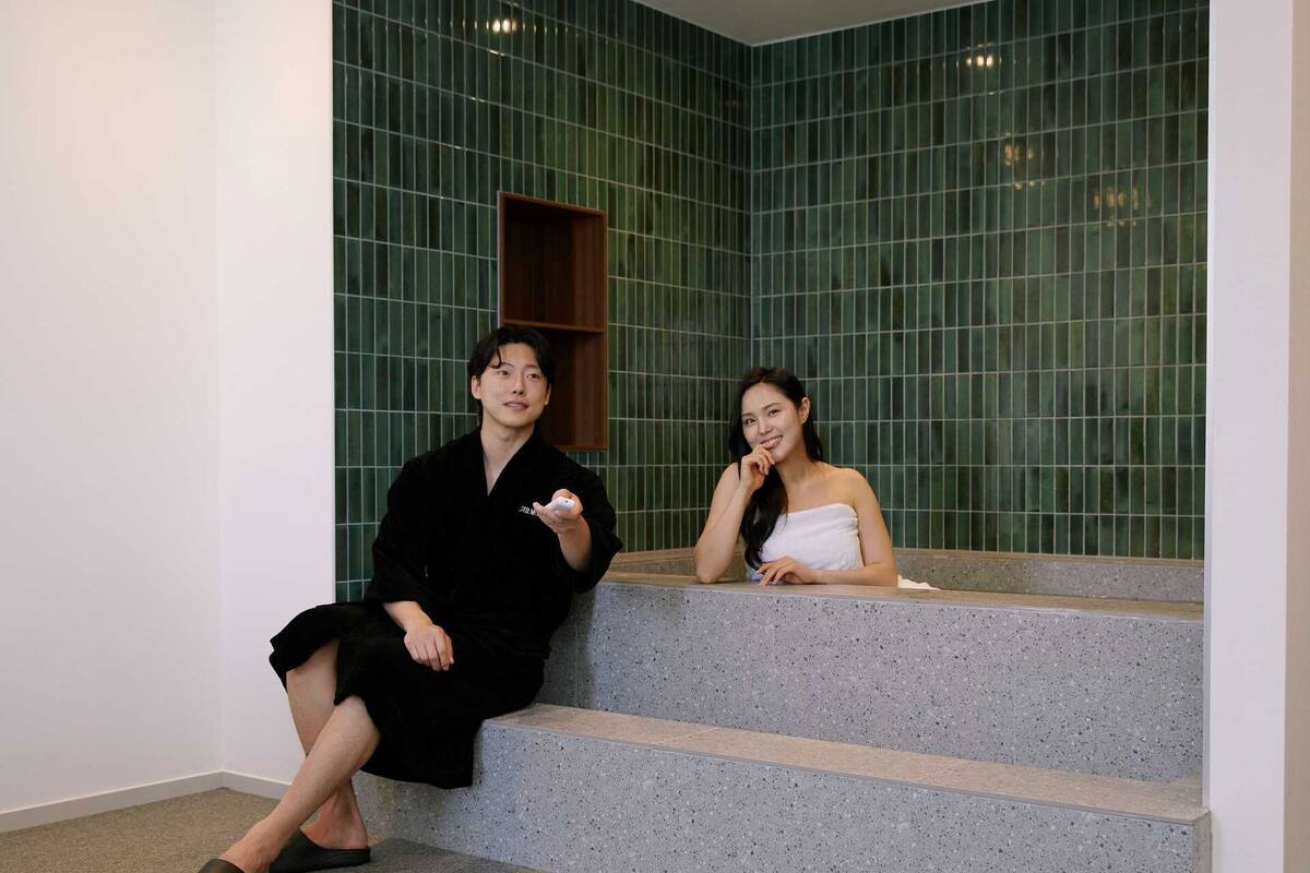 Two individuals seated on a stone bench in a room with green tiled walls and a wooden shelf.