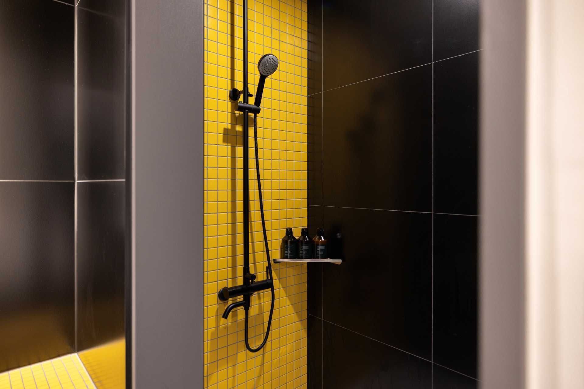 A shower with a yellow tiled wall, a handheld showerhead, and a black hose.