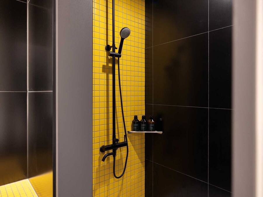 A shower with a yellow tiled wall, a handheld showerhead, and a black hose.
