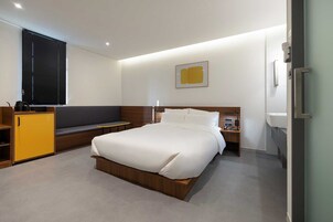 Una habitación de hotel moderna con una cama grande, una zona de estar con un banco, un escritorio pequeño y una ventana con persianas.
