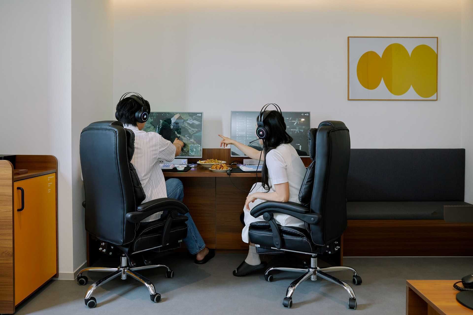 Duas pessoas sentadas à mesa com computadores, fones de ouvido e um prato de comida.