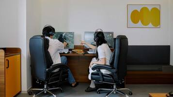 Duas pessoas sentadas à mesa com computadores, fones de ouvido e um prato de comida.