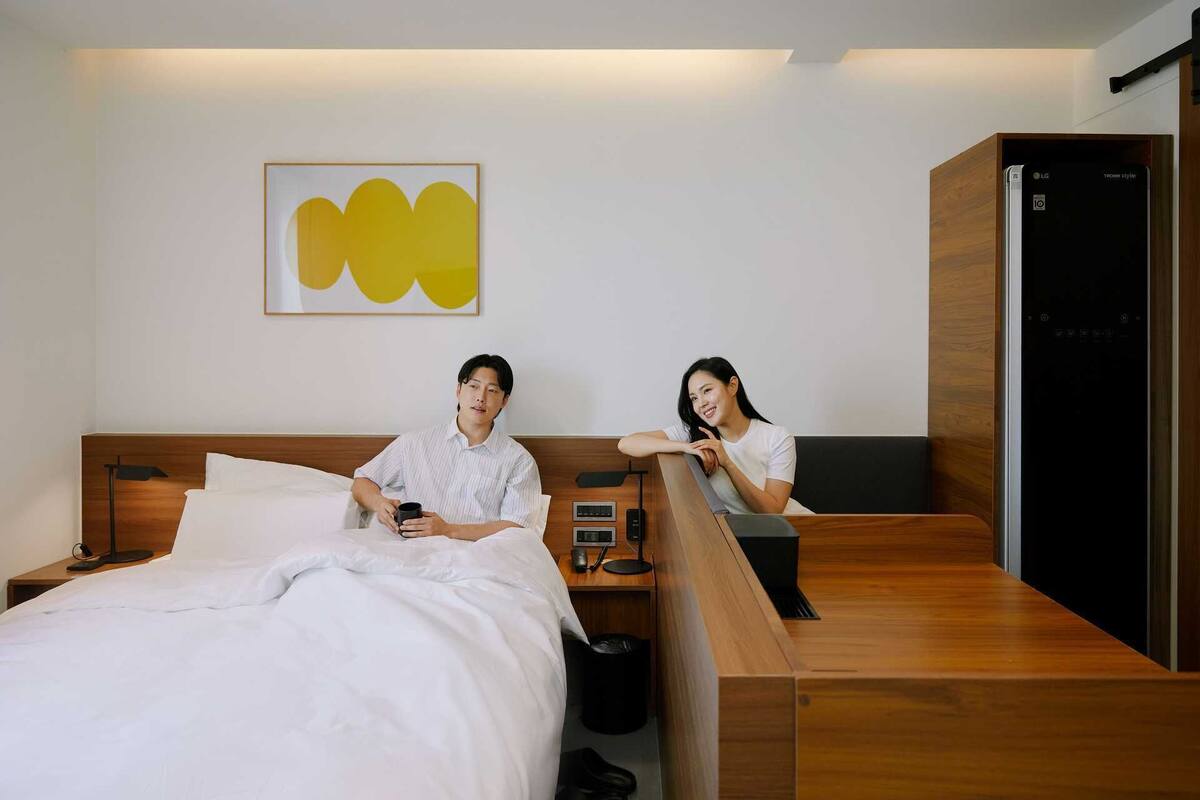 A modern hotel room with a wooden headboard, a bed with white linens, a bedside table with a lamp, a small trash bin, and a wooden bench.
