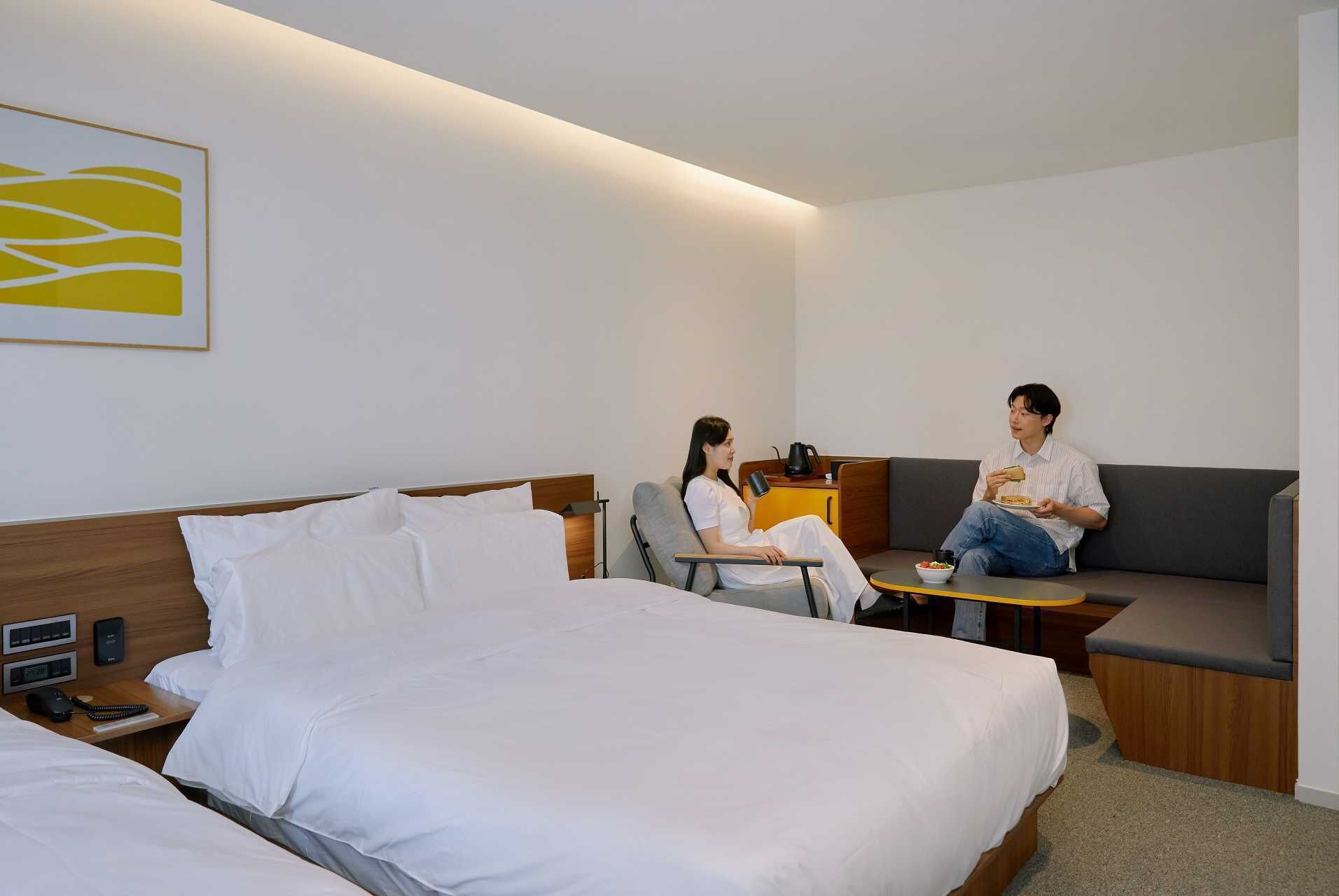 A modern hotel room with a large bed, a sofa, and a small table. There is a painting on the wall and a coffee maker on the table.