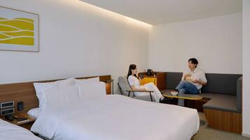 A modern hotel room with a large bed, a sofa, and a small table. There is a painting on the wall and a coffee maker on the table.