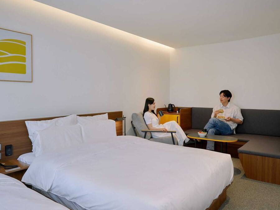A modern hotel room with a large bed, a sofa, and a small table. There is a painting on the wall and a coffee maker on the table.