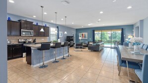 A modern kitchen with dark cabinets, a central island with bar stools, and a dining area with a large wooden table and blue chairs. - 8BR Villa! Free Arcade, Pool, Spa Windsor Westside (Kissimmee)