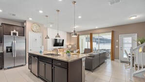 A modern kitchen with a central island, stainless steel appliances, pendant lighting, and a view to the living area.