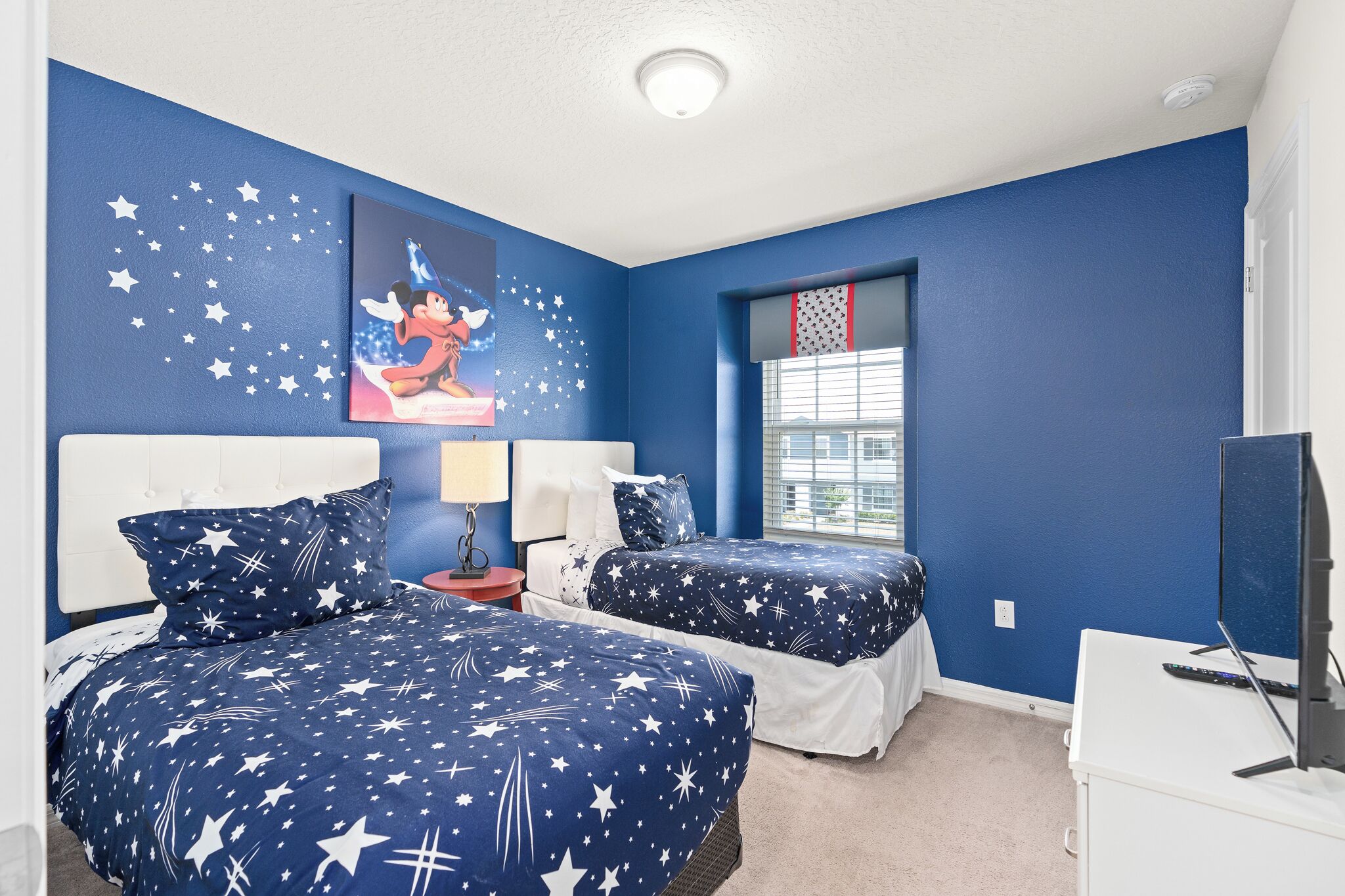 Een slaapkamer met twee bedden, een nachtkastje, een lamp, een televisie en een raam met jaloezieën.