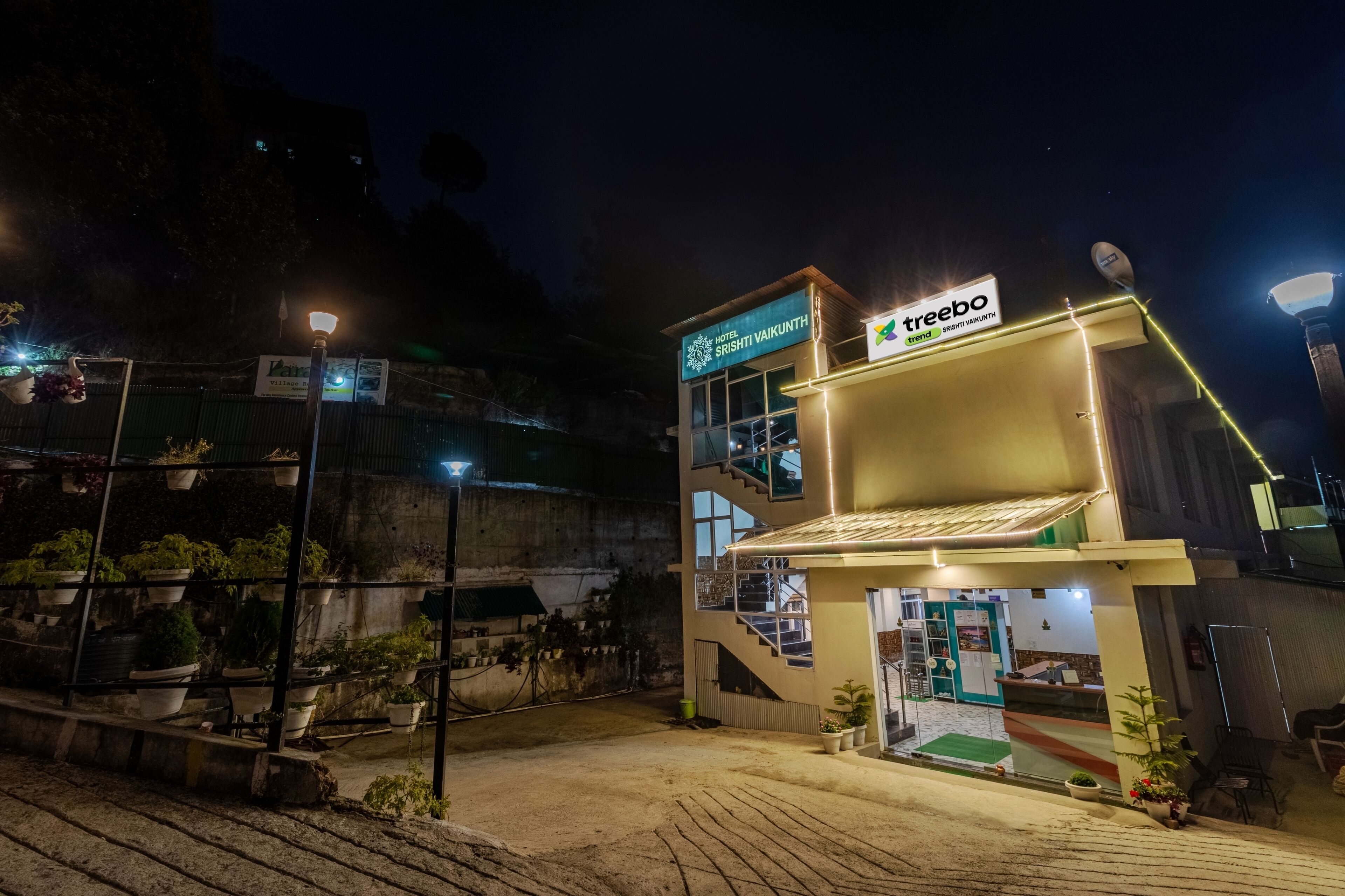 A hotel named 'Hotel Srishti Vaikunth' with a signboard, a staircase leading to the entrance, and a small outdoor seating area with potted plants.